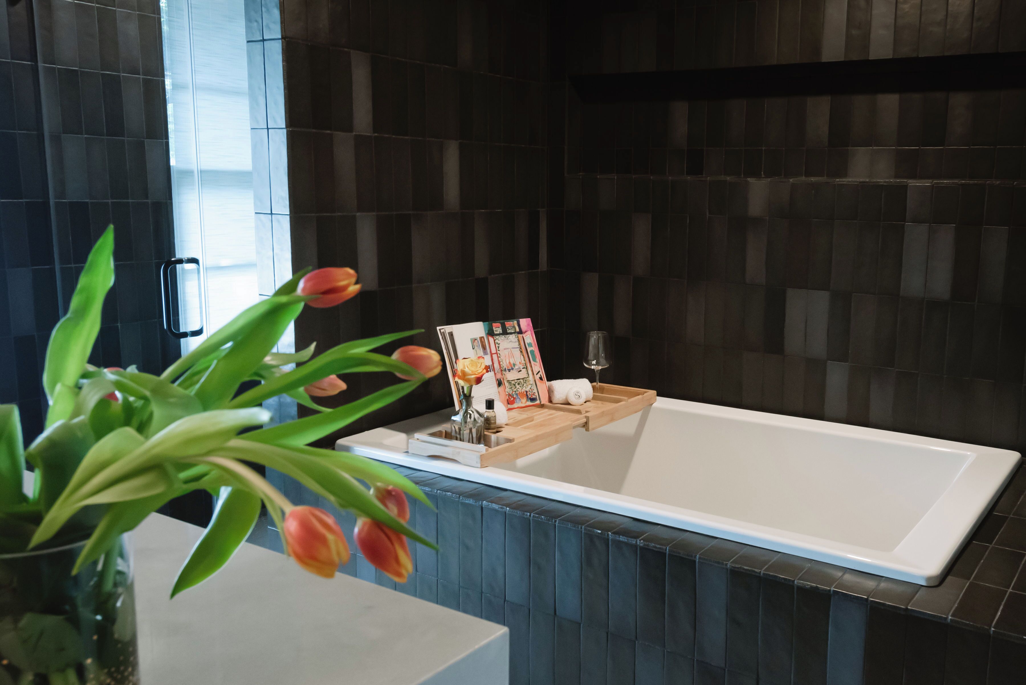 Une salle de bain moderne avec une grande baignoire, des murs recouverts de carreaux foncés, un plateau en bois contenant des articles de toilette et un vase rempli de tulipes.