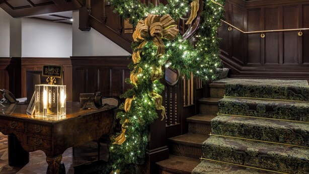 A grand staircase with a green garland and golden bows, flanked by wooden paneling and framed artwork on the walls. - La Colombe d'Or Hotel (Houston)