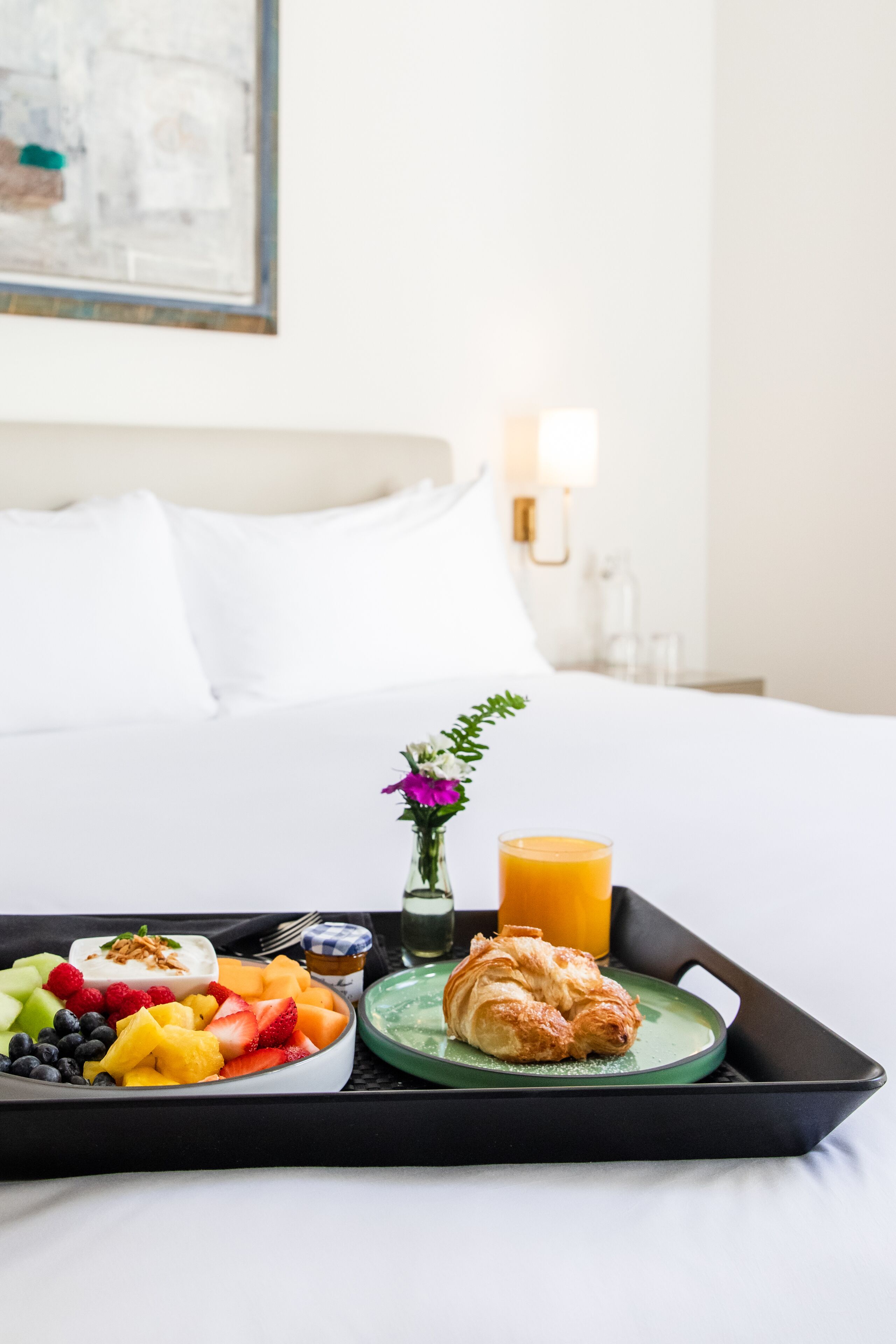 A breakfast tray with fruits, yogurt, a croissant, and orange juice on a bed in a hotel room.