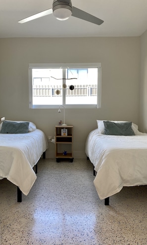 A bedroom with two beds, a ceiling fan, a window with a view of a fence, and a small bedside table with a lamp.