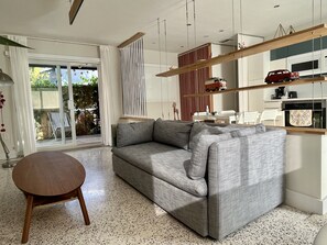 Una sala de estar moderna con un sofá gris, una mesa redonda de madera y una cocina visible a través de un vidrio.