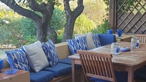 A wooden table and bench set with blue cushions, surrounded by greenery and a wooden lattice structure. - Costa Serena, cottage near the sea. (Palau)