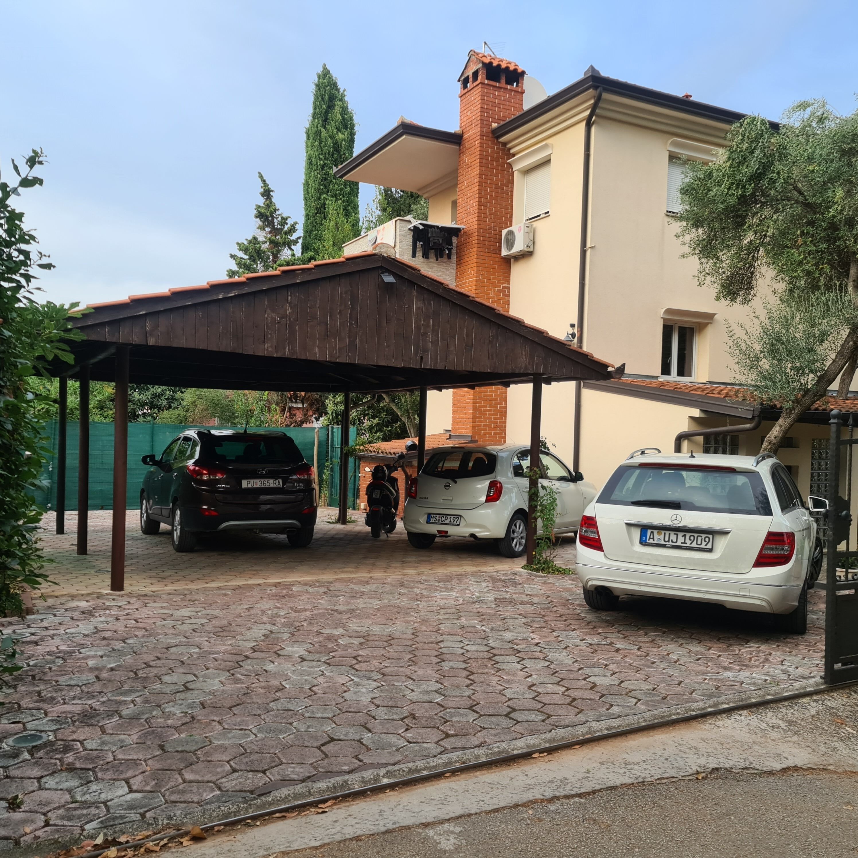 A covered parking area with three cars parked, a brick building with a chimney, and a green fence in the background.