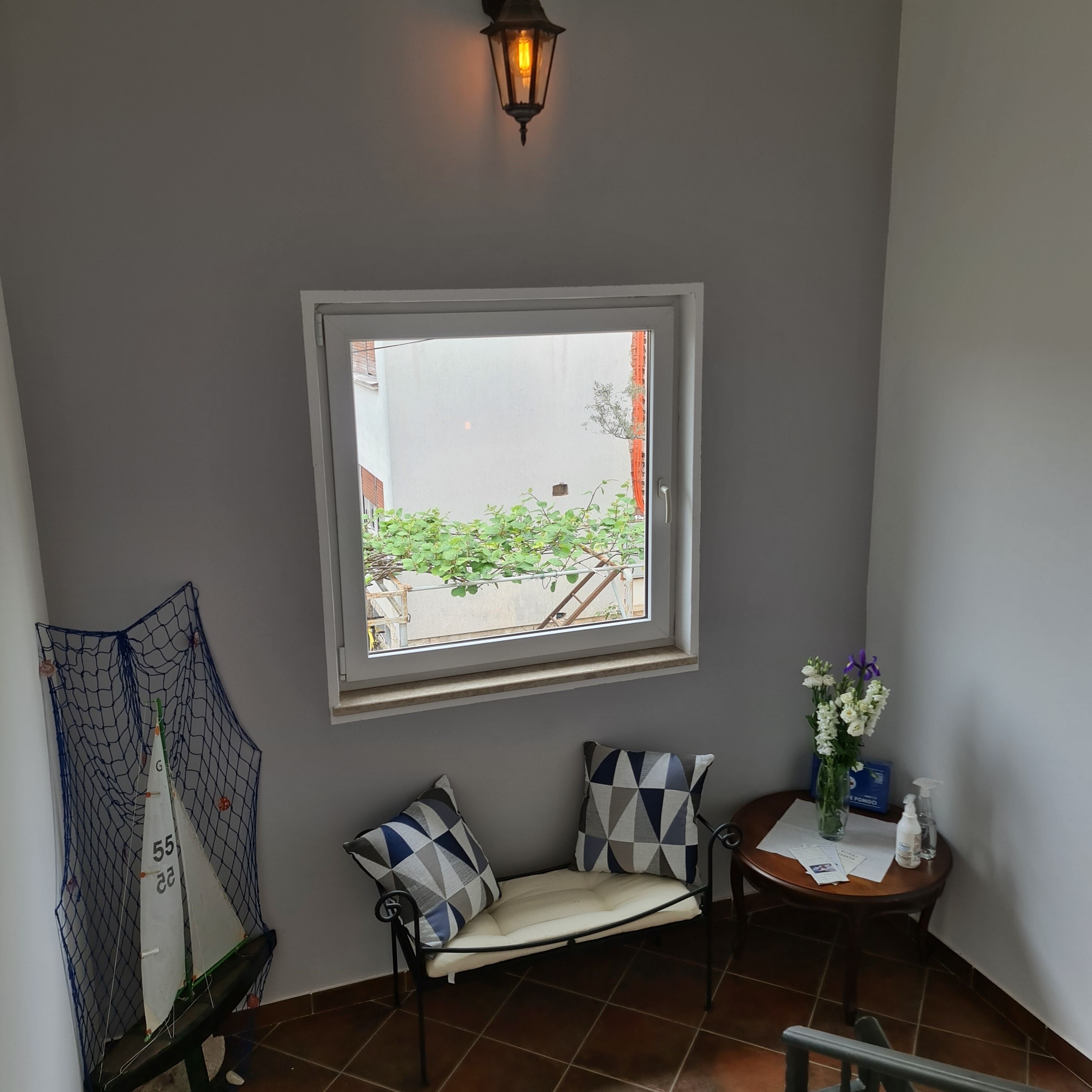 Habitación de esquina con ventana, un banco con cojines estampados, una mesita con un florero y una lámpara fijada a la pared.