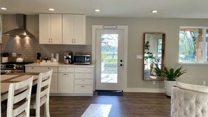 Cocina moderna con gabinetes blancos, estufa de acero inoxidable y microondas. Hay un comedor con mesa y sillas de madera, y un espejo grande en la pared.