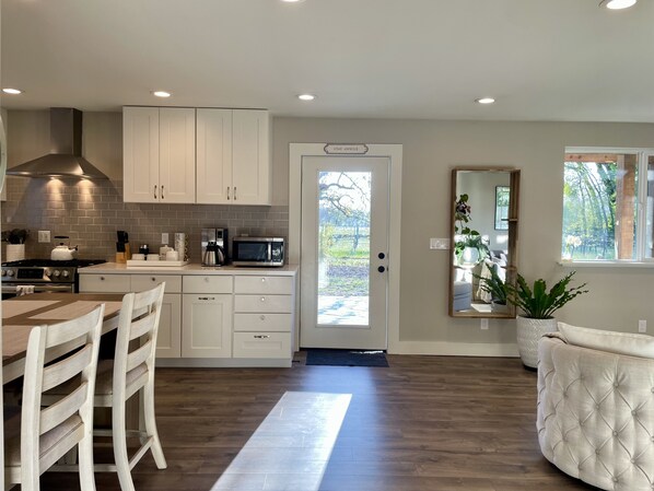 A modern kitchen with white cabinets, a stainless steel range, and a microwave. There's a dining area with a wooden table and chairs, and a large mirror on the wall.