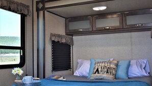 Een slaapkamer met een bed, een raam met uitzicht, een nachtkastje met een kopje en boeken, en een hoofdbord met een decoratief kussen.