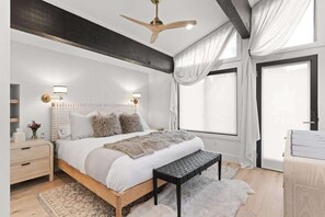Un dormitorio con cama, mesitas de noche, ventilador de techo, ventana con cortinas y vistas al exterior.