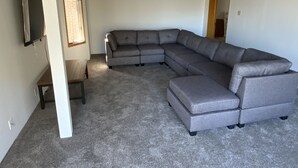 A living room with a large L-shaped sectional sofa, a flat-screen TV mounted on the wall, and a window with blinds. - BEACHVIEW GETAWAY (Sheboygan)