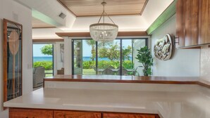 A modern kitchen with a wooden countertop, a hanging chandelier, and a view of the ocean through large windows. - Oceanfront luxury 3bd condo on Baby Beach (Lahaina)