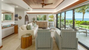 A modern living room with a white sofa, grey armchairs, a wooden coffee table, and a large window offering a view of the ocean. - Oceanfront luxury 3bd condo on Baby Beach (Lahaina)