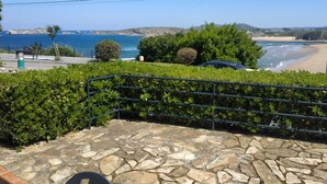 Un balcon dallé de pierres, doté d’une rambarde en métal, qui offre une vue sur une zone côtière avec une plage et des falaises rocheuses.