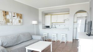 Un salon moderne avec un canapé gris, une table blanche et un coin cuisine doté de meubles blancs et d’un dosseret carrelé.