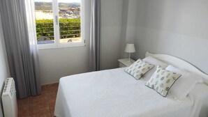 Une chambre à coucher avec un lit, une fenêtre donnant sur un paysage verdoyant et des bâtiments, une table de chevet avec une lampe et un radiateur.