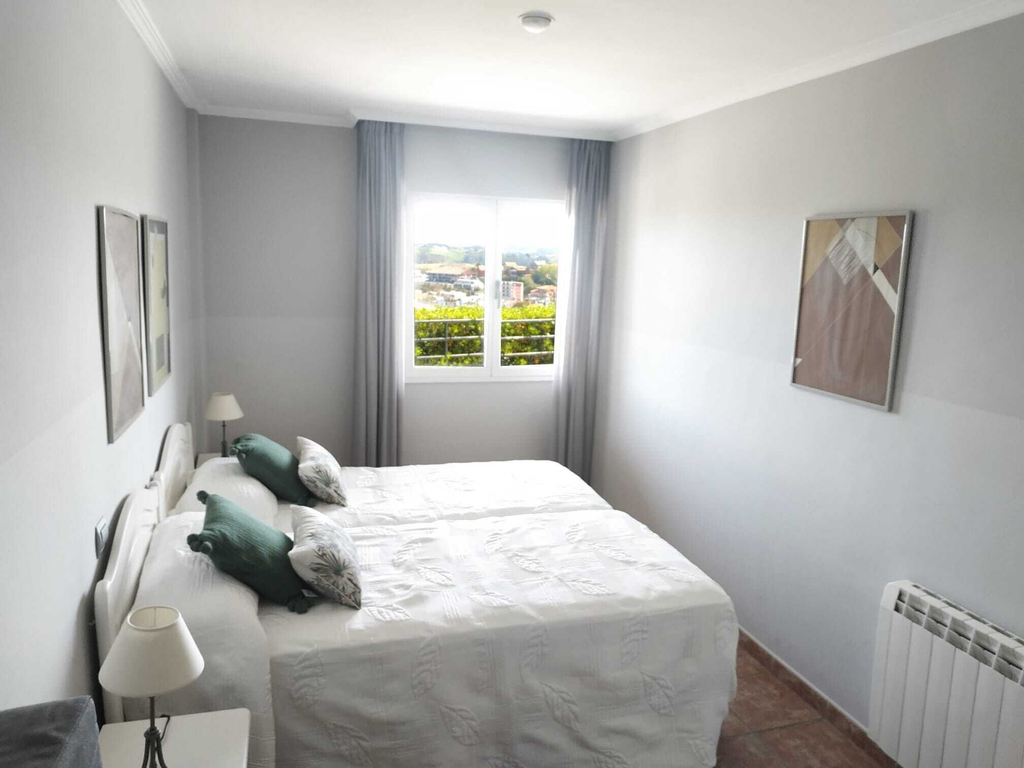 Un dormitorio con una cama grande, dos almohadas, una mesita de noche con lámpara, una ventana con cortinas y un radiador.
