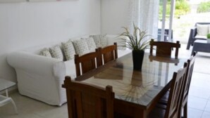 Een eetruimte met een tafel met glazen blad, houten stoelen, een witte bank met gekleurde kussens en een potplant.