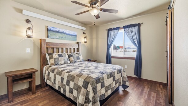 A bedroom with a wooden bed, a ceiling fan, a nightstand, a window with curtains, and a painting on the wall.
