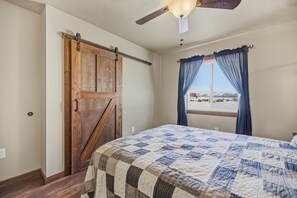 Een slaapkamer met een groot bed, een houten deur in boerderijstijl, een plafondventilator en een raam met gordijnen.