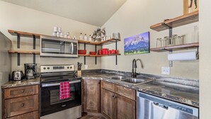 A kitchen with wooden cabinets, a stainless steel dishwasher, a microwave, and a stove. There are shelves above the stove holding jars and a painting on the wall.