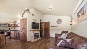 A spacious living area with a fireplace, a brown leather couch, wooden flooring, a kitchen with modern appliances, and a mounted TV.
