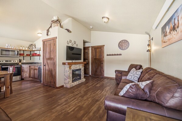 A spacious living area with a fireplace, a brown leather couch, wooden flooring, a kitchen with modern appliances, and a mounted TV. - 2BR Family Cabin Near Yellowstone (Island Park)