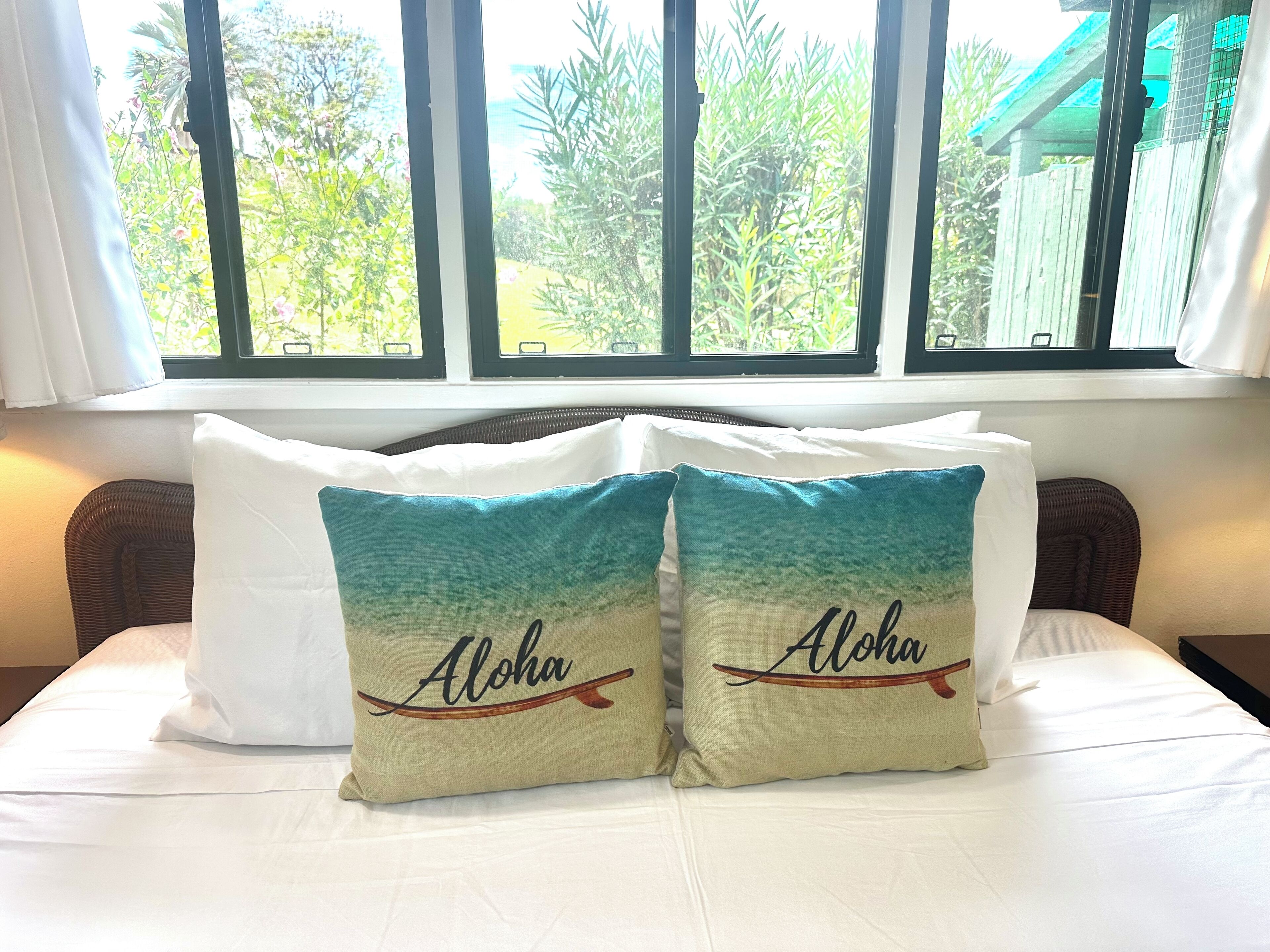 A bed with white bedding and two pillows with 'Aloha' text and a wave design.