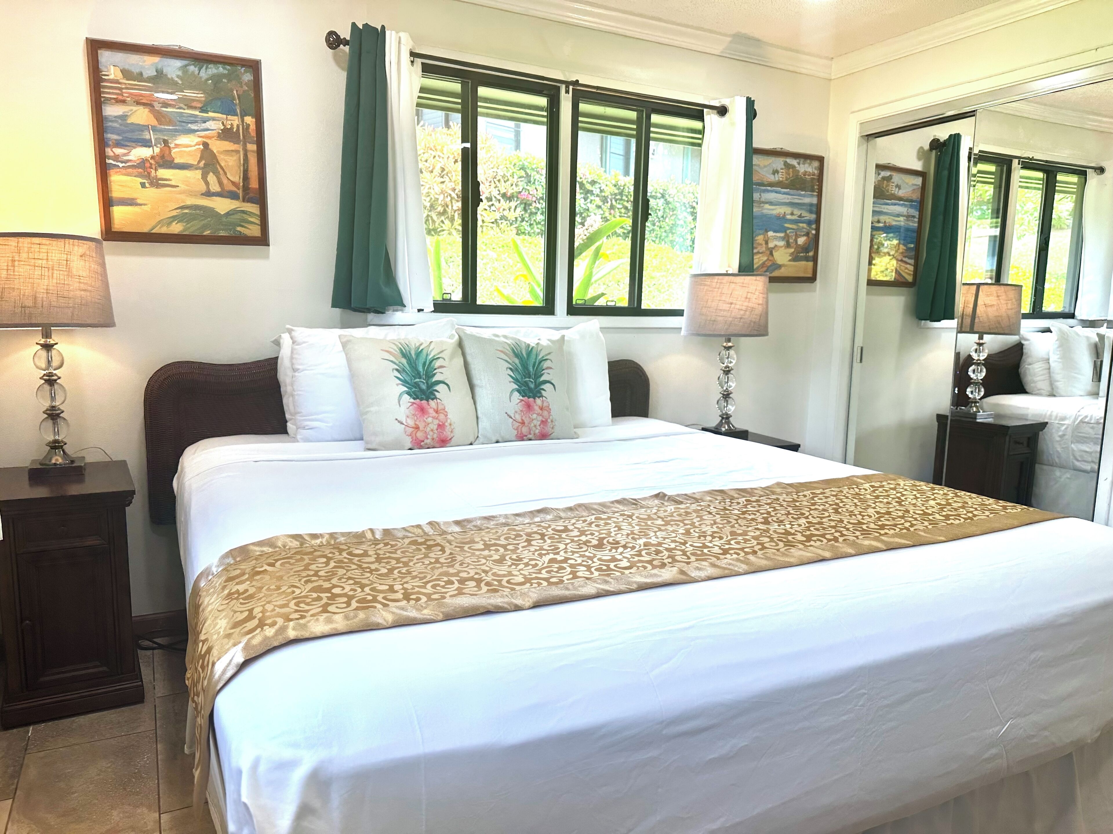 Una cama bien hecha con un edredón estampado, dos almohadas con diseño de piña y una ventana con cortinas verdes.