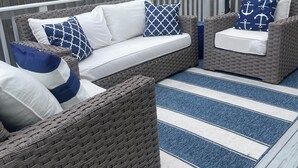 Un espace de détente extérieur aménagé avec des meubles en osier, un tapis à rayures bleues et blanches, des coussins blancs et un parasol blanc.