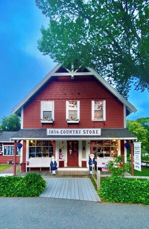 A red building labeled "1856 Country Store" with a porch and a statue of a man. - WALK TO THE BEACH LUXURY VACATION CENTRAL AIR, KG BEDS, IDEAL FOR LONG STAYS. (Barnstable)
