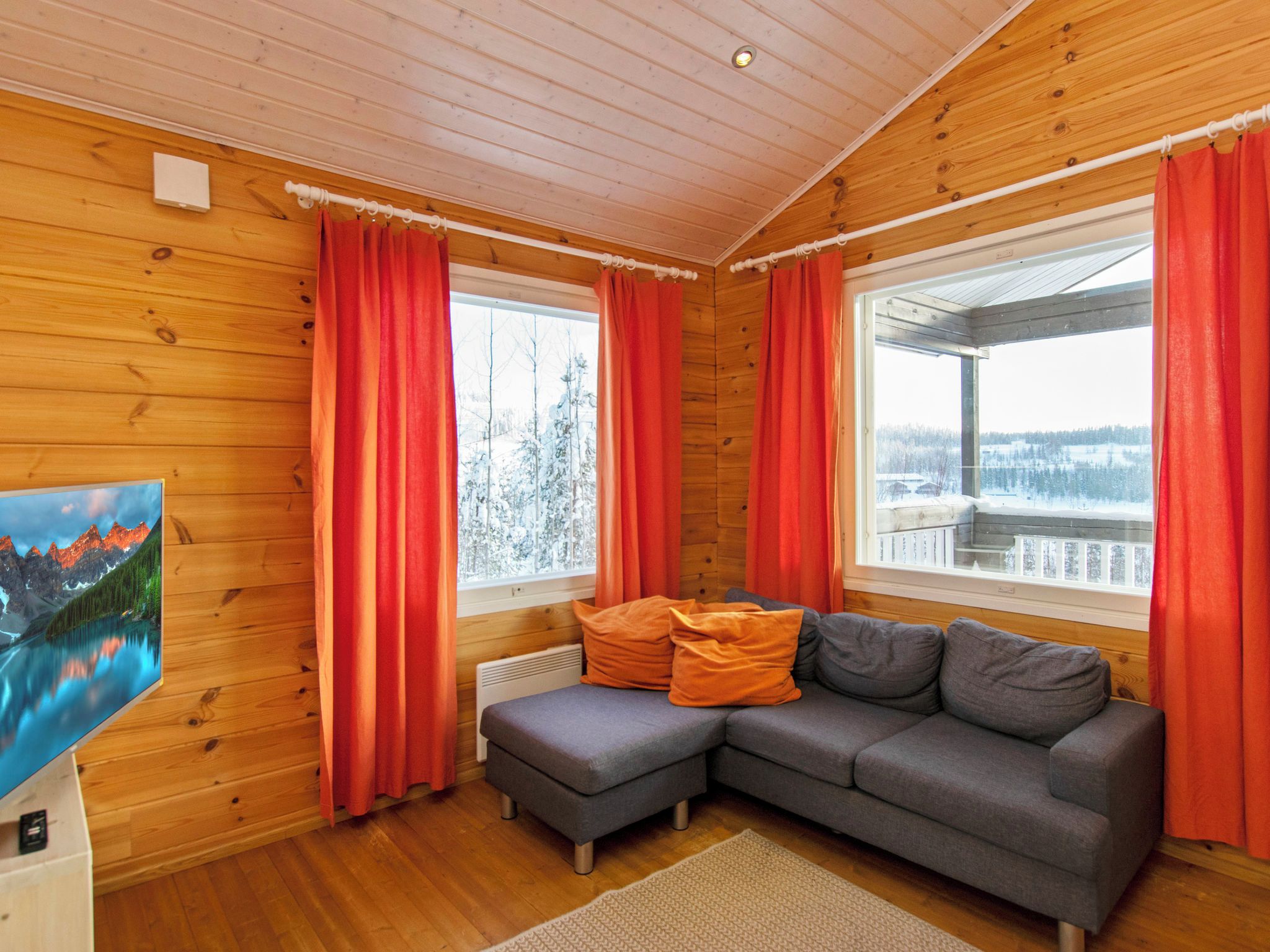A cozy cabin interior with a sectional sofa, a television, and large windows with red curtains.