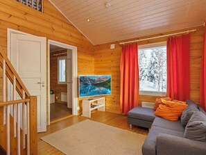 A cozy cabin interior with a wooden interior, a television on a stand, a gray sofa, and a window with red curtains.