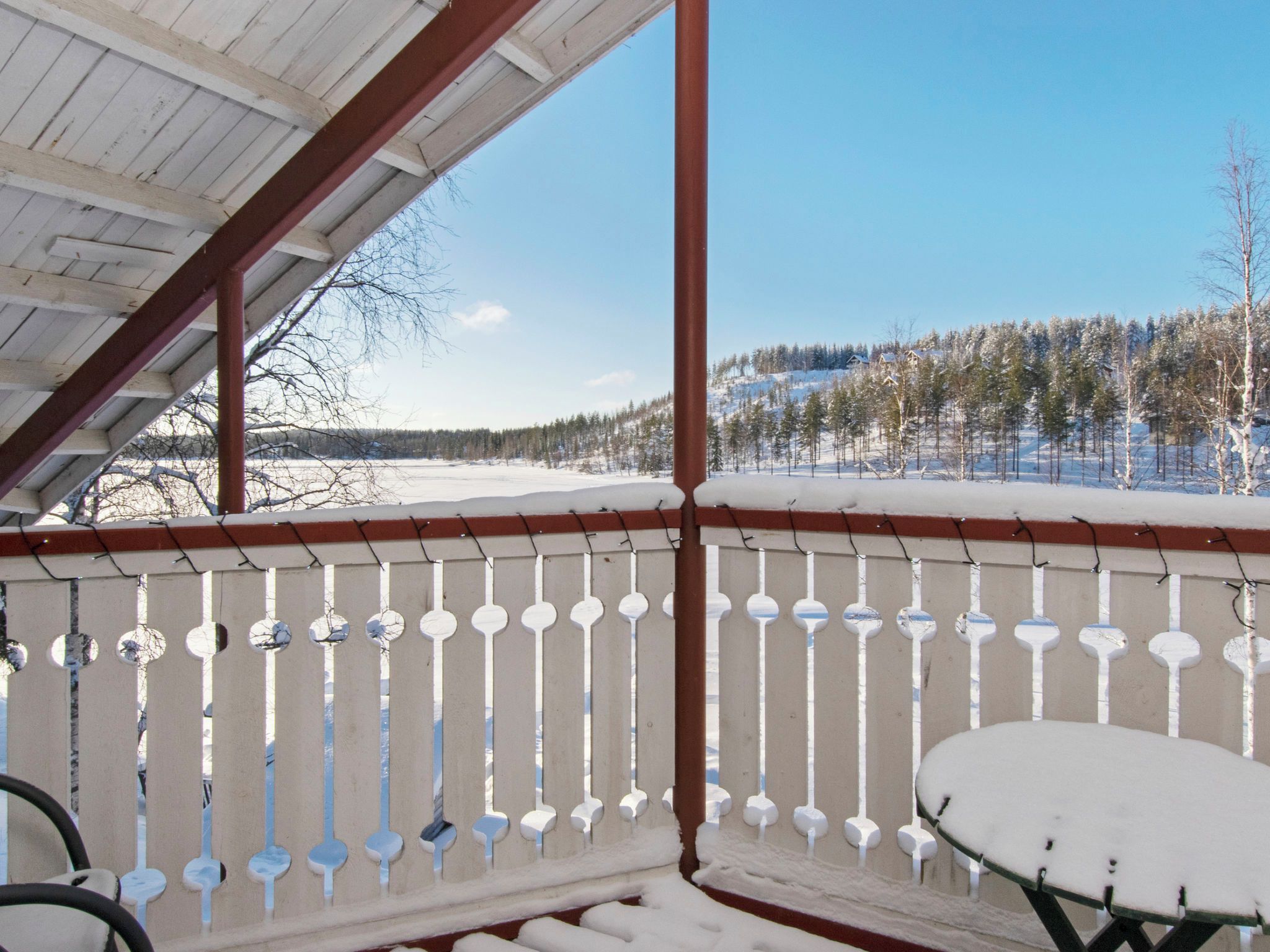 Un espace extérieur abrité, avec une table et des chaises recouvertes de neige, une clôture en bois et une vue sur un paysage enneigé, avec des arbres et une maison au loin.