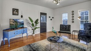 Un salon moderne avec un plancher en bois, un meuble bleu, un téléviseur, une grande plante, une statue de paon, un fauteuil noir, une table ronde et un ventilateur de plafond.