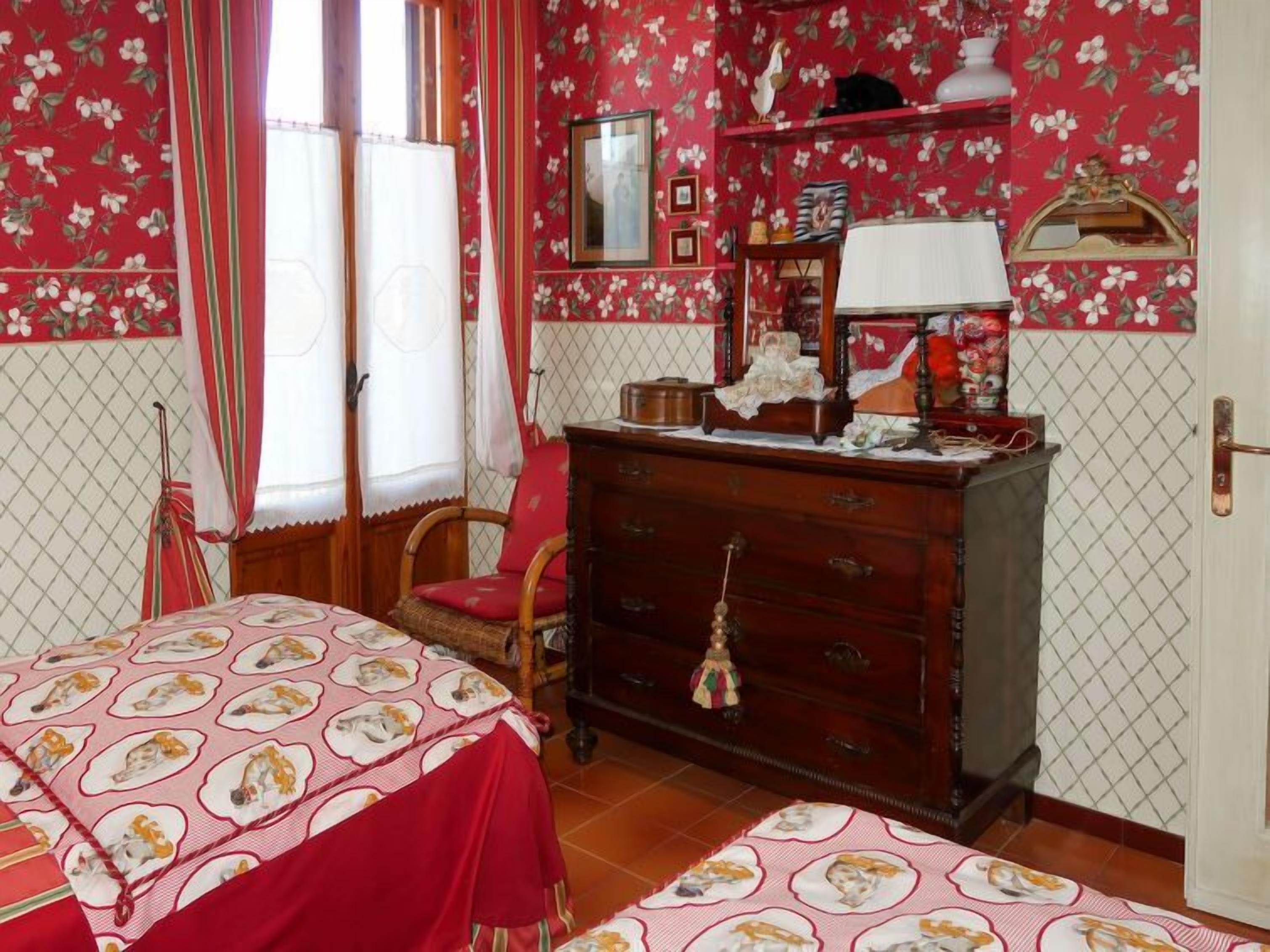 A room with floral wallpaper, a wooden dresser, a bed with a red bedspread, and a chair.