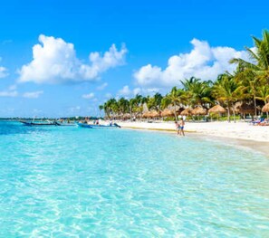 A beach with clear turquoise water, palm trees, and a few boats.