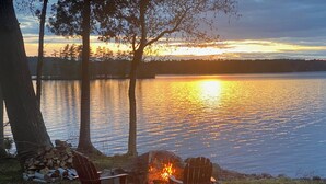 A lakeside setting with a fire pit and two chairs, surrounded by trees and a view of the sunset over the water. - Stunning Private Lakehouse with Dock and outdoor Hot Tub (Manchester)