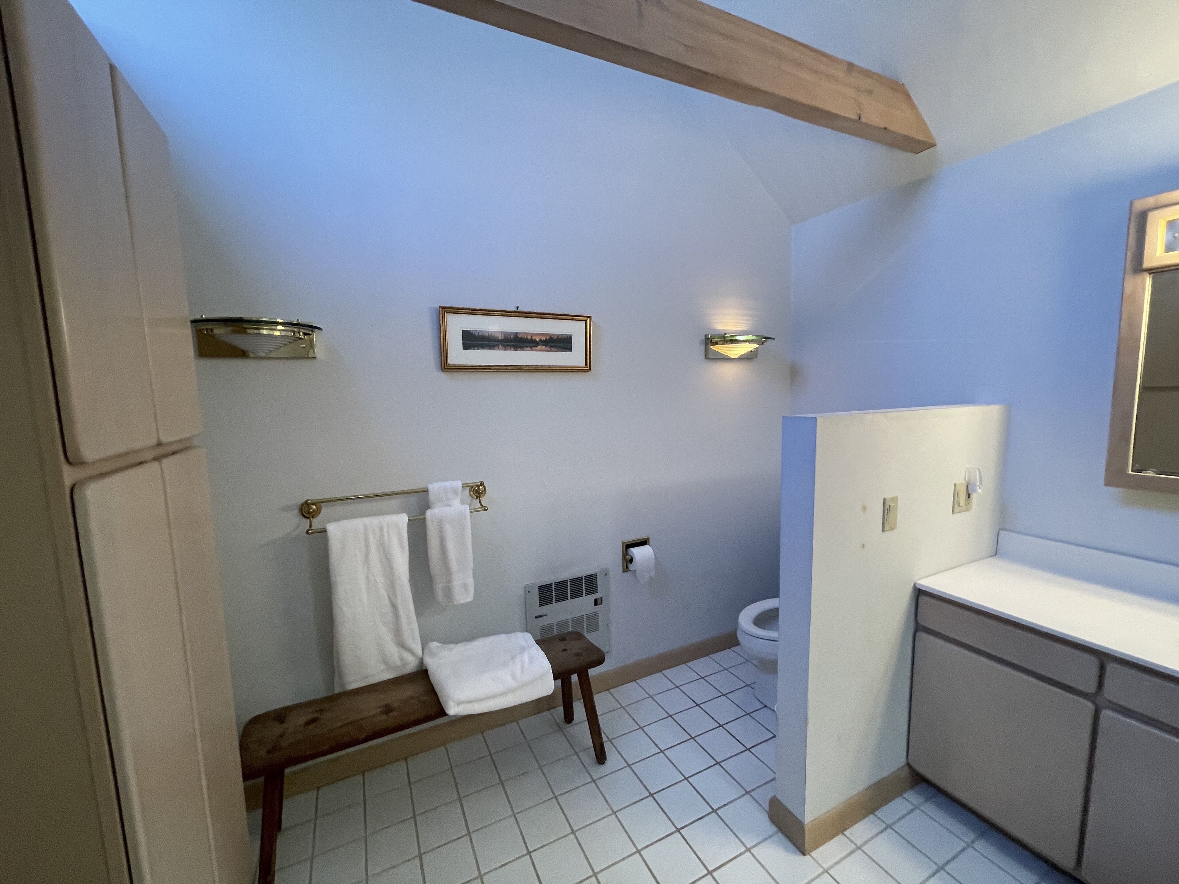 Un baño con banco de madera, inodoro, lavamanos y un cuadro en la pared.