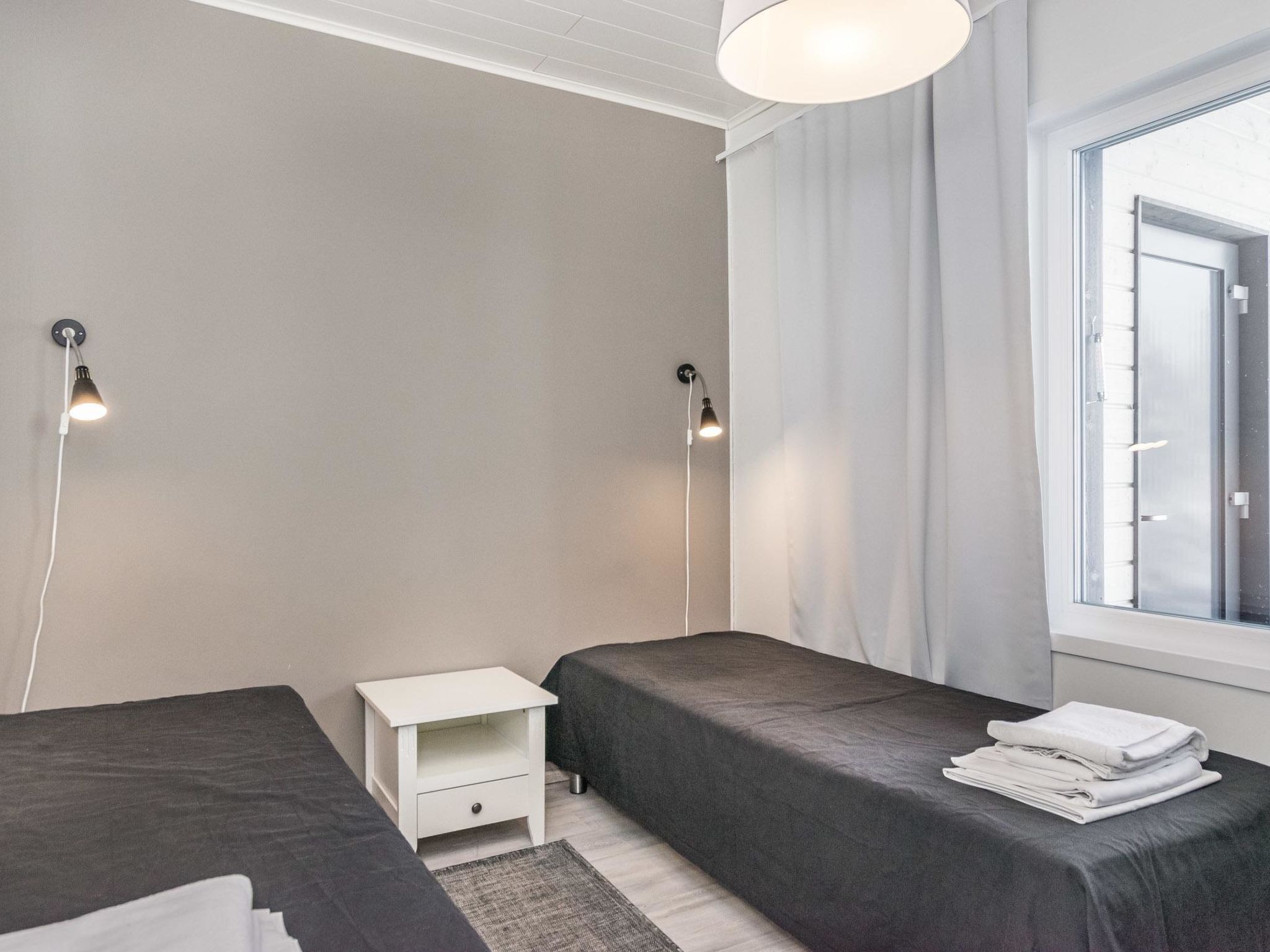 A modern bedroom with two beds, a small white nightstand, and a window with curtains.