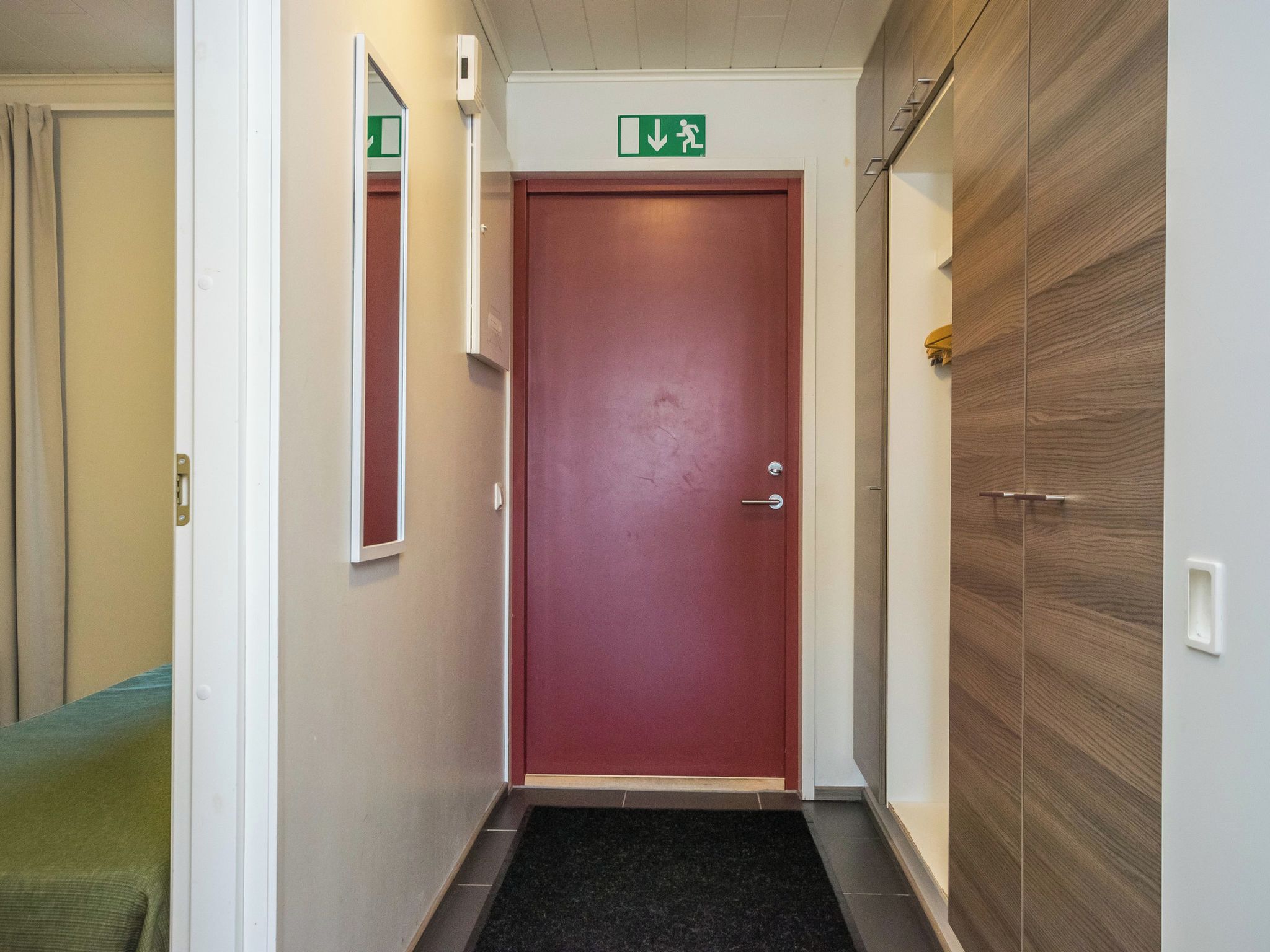 A hallway with a red door, a green exit sign, and a wooden wardrobe.