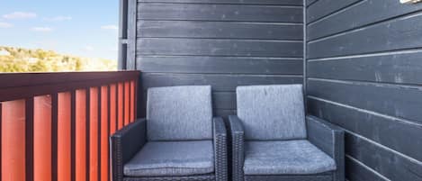 Twee rieten stoelen met grijze kussens op een houten terras, naast een donkere houten wand.