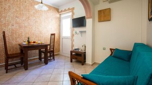 A living room with a brick wall, a dining table with chairs, a teal sofa, a TV mounted on the wall, and an air conditioning unit. - Gardenia Eco Apartment in Panormo (Panormos)