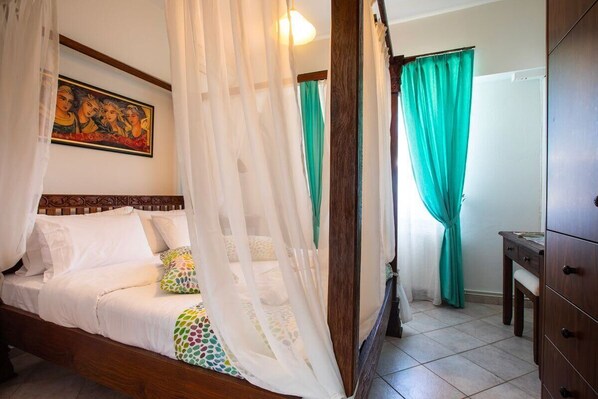 Una cama con cabecera de madera, dosel y almohadas coloridas. Hay un cuadro en la pared y una cortina verde.