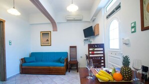 Ein Wohnzimmer mit einem blauen Sofa, einem Esstisch mit Obst und Gläsern sowie einem Fernseher an der Wand.
