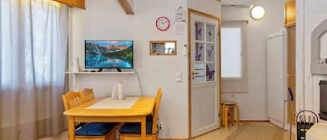 A cozy room with a wooden dining table, chairs, a flat-screen TV, a clock, and a window with curtains.
