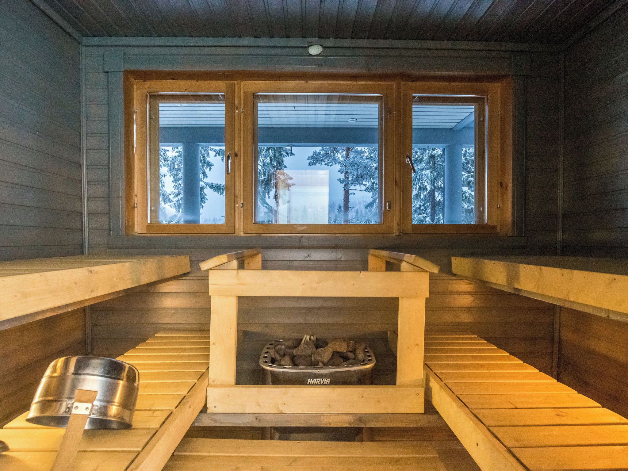 A sauna room with a wooden bench, a metal container, and a Harvia wood-burning stove.