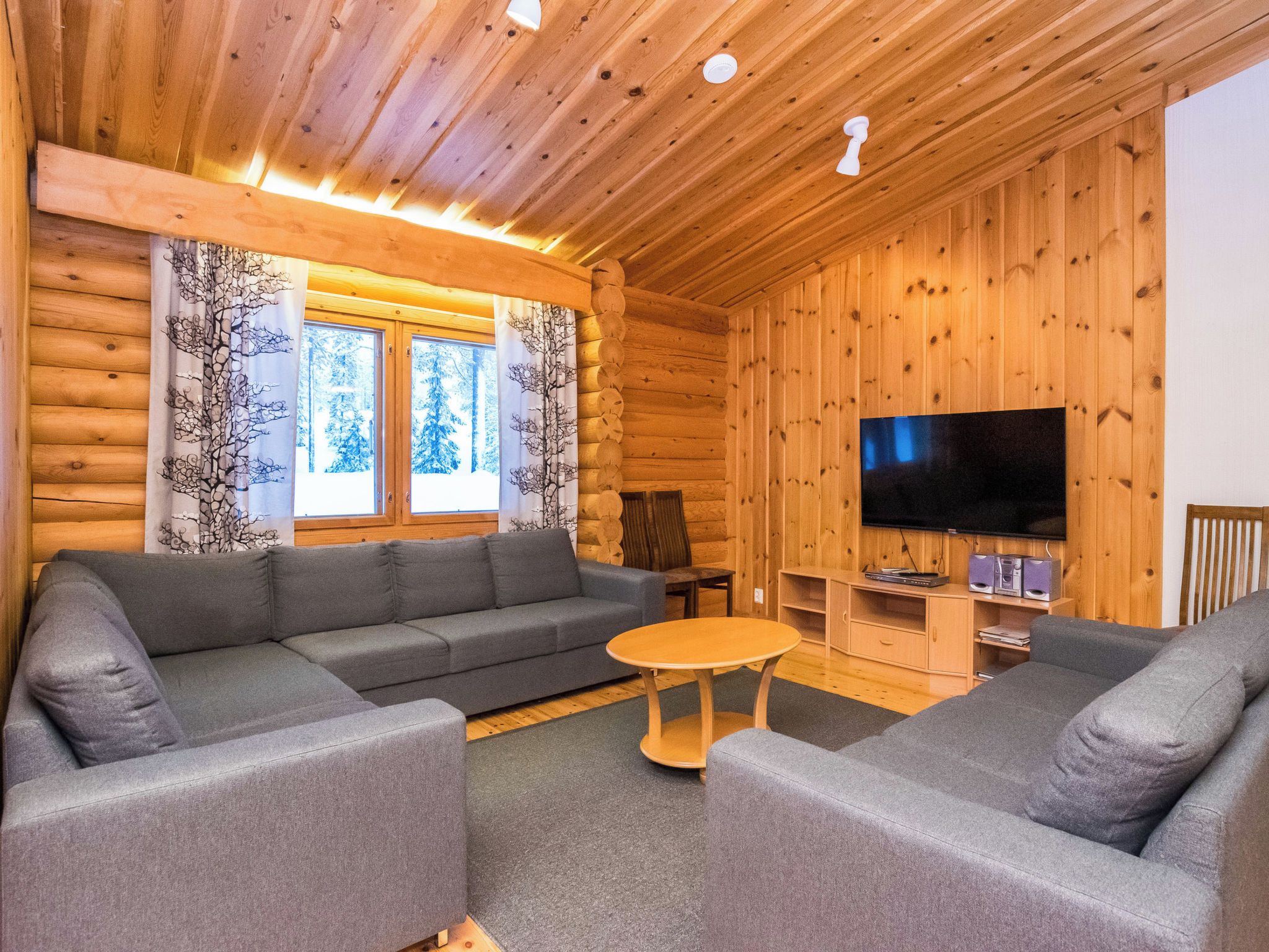 L’intérieur d’une cabane en rondins, avec un canapé gris, une petite table ronde et un téléviseur à écran plat fixé au mur.