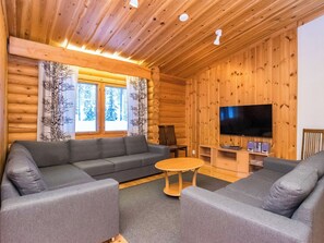 L’intérieur d’une cabane en rondins, avec un canapé gris, une petite table ronde et un téléviseur à écran plat fixé au mur.