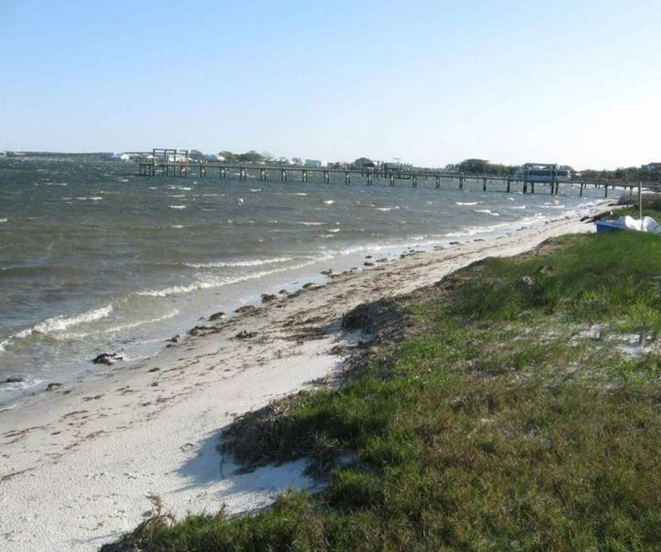 Una playa con un muelle que se adentra en el agua, una orilla arenosa y zonas verdes.