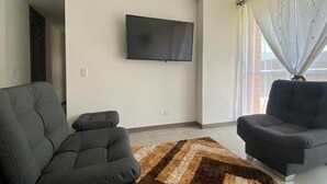 A living room with a flat-screen TV mounted on the wall, two dark sofas, and a patterned rug.