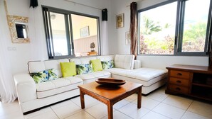 Uma sala de estar moderna com um sofá branco, uma mesa de centro de madeira e amplas janelas com vista para o verde.